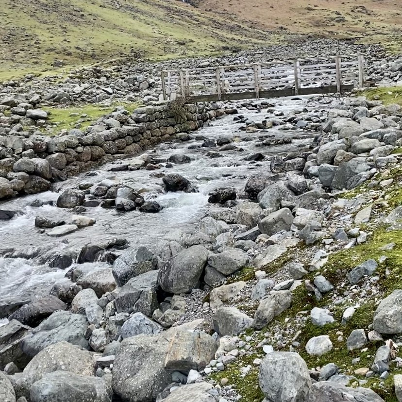 A river lined with boulders and a wooden bridge crossing it.