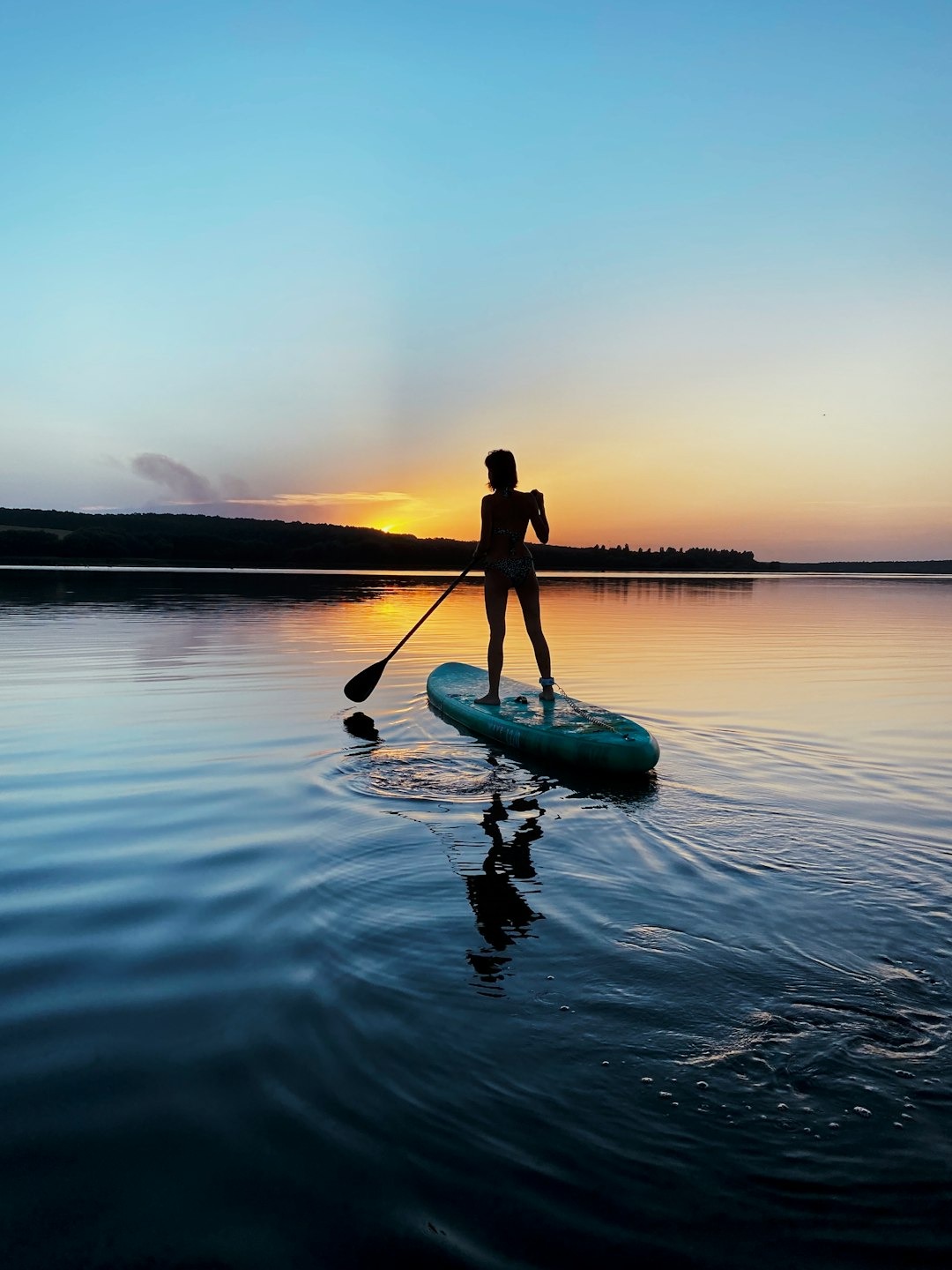 A person stand up paddle boarding on a lake with the sunset behind them.
