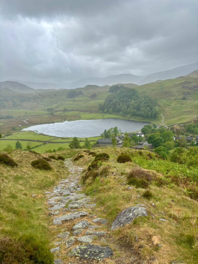 A view of Watendlath Tarn, a paved path and the surrounding trees and fells