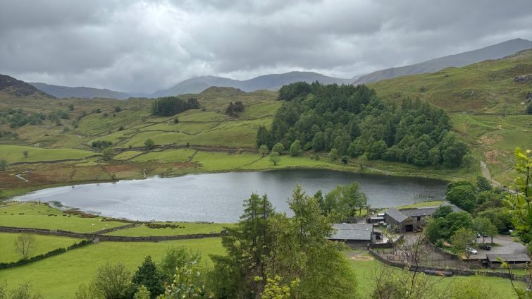 A view of Watlendlath Tarn, Watlendlath, in the Lake District. Lush green rolling hills and trees surround the tarn.
