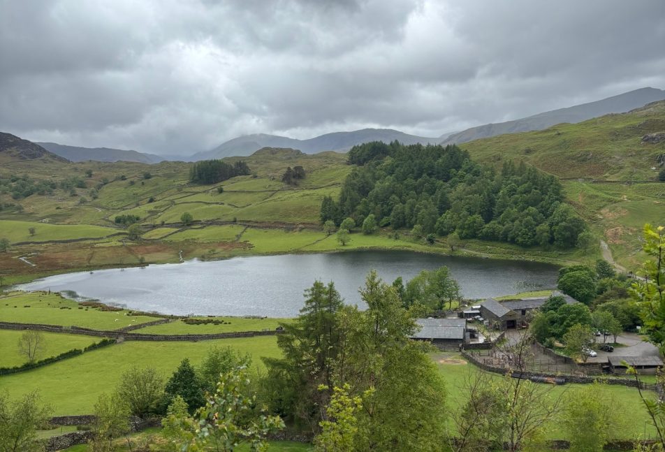A view of Watlendlath Tarn, Watlendlath, in the Lake District. Lush green rolling hills and trees surround the tarn.